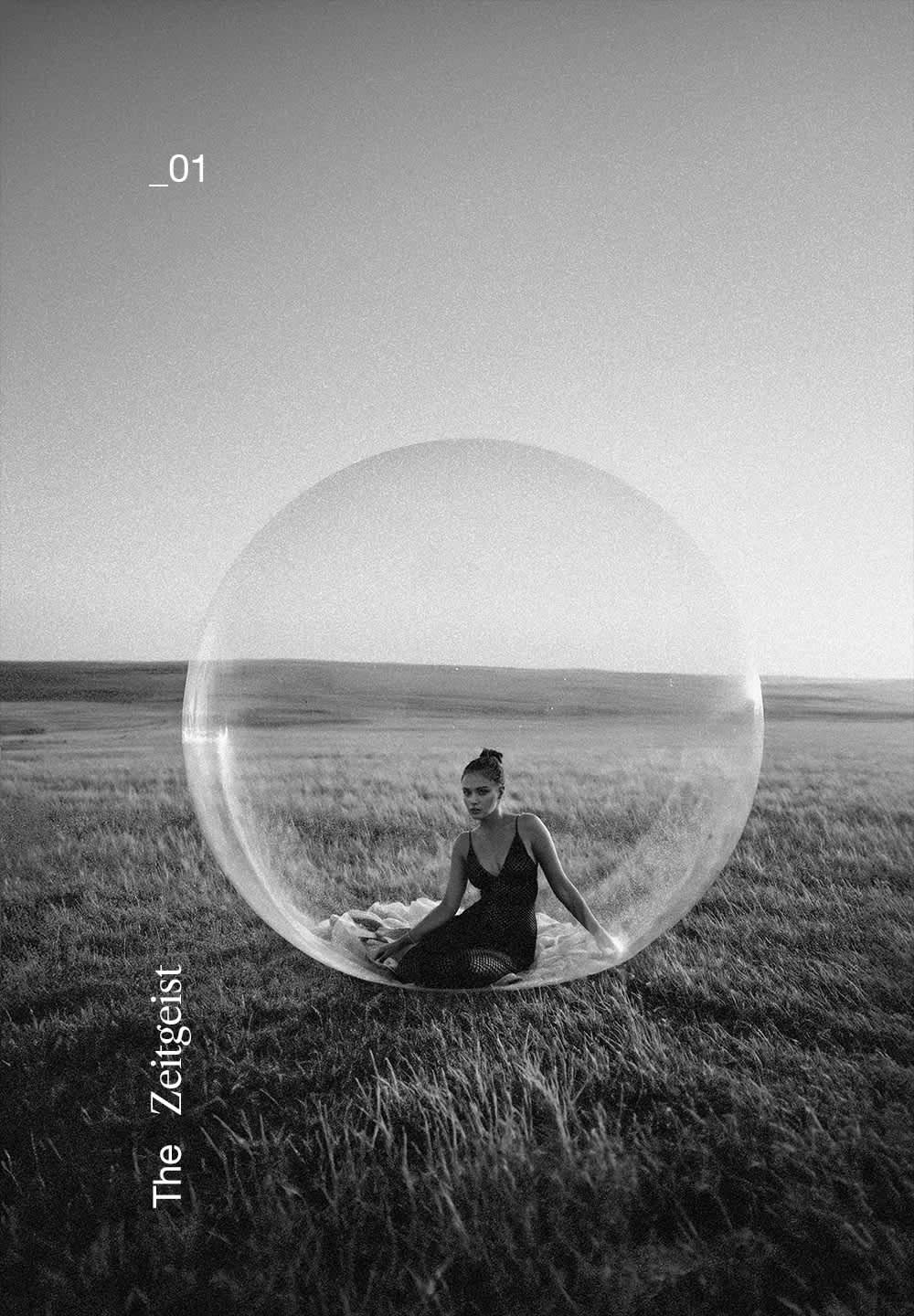 Clear plastic bubble structure on grassy field, with a figure seated on a blanket inside. Black clothing contrasts with the transparent surface.