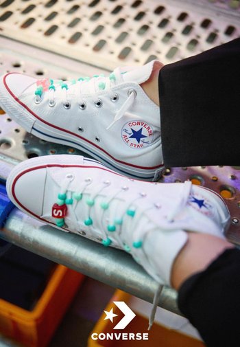 Baskets blanches Converse All Star avec lacets turquoise et détail perle rouge portées par une personne assise sur une surface métallique.