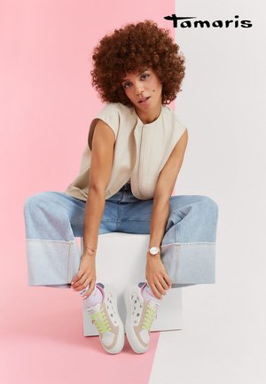 Woman with curly hair sits on white block against pink and white background, wearing sleeveless beige top, wide-leg jeans, and floral sneakers.