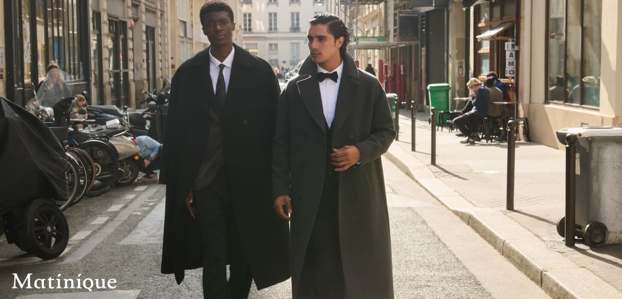 Zwei Männer gehen eine Stadtstraße entlang, sie tragen lange dunkle Mäntel. Die Szene zeigt städtische Architektur, Fahrräder und Sitzgelegenheiten in einem Straßencafé.