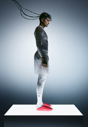 Een persoon in grijze sportkleding staat op een wit platform, draagt rode Nike-schoenen met textuurdetails. Lang, donker haar stroomt naar achteren.