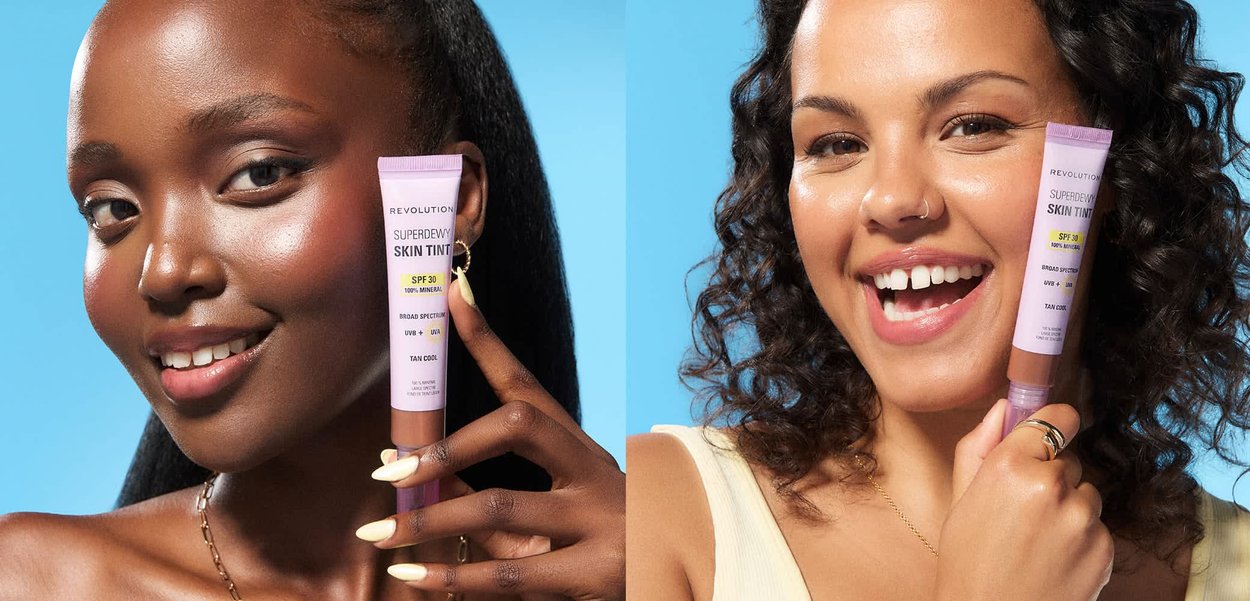 Due donne sorridenti con tonalità di pelle diverse tengono in mano tubetti di Revolution Superdew Skin Tint SPF 30 su uno sfondo blu.