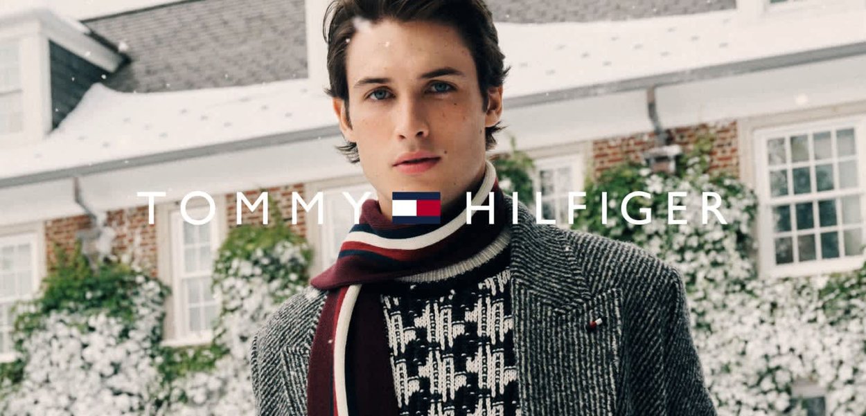 Cappotto di lana grigio con motivo strutturato, indossato sopra un maglione lavorato a maglia bianco e nero. Sciarpa a righe rosse e blu navy avvolta intorno al collo. Sfondo innevato.