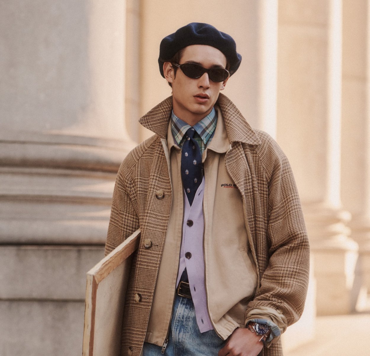 Jonge man met een geruite jas, beige jas, lavendelkleurig vest, geruit overhemd, marineblauwe stropdas, zwarte baret en zonnebril, die een canvasframe vasthoudt.