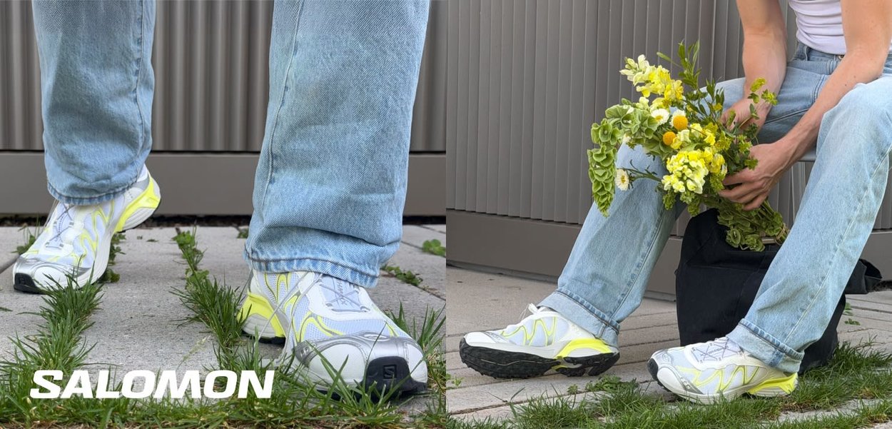 Person iført lyseblå jeans og hvide Salomon-sneakers står og sidder udendørs og holder en buket gule blomster.