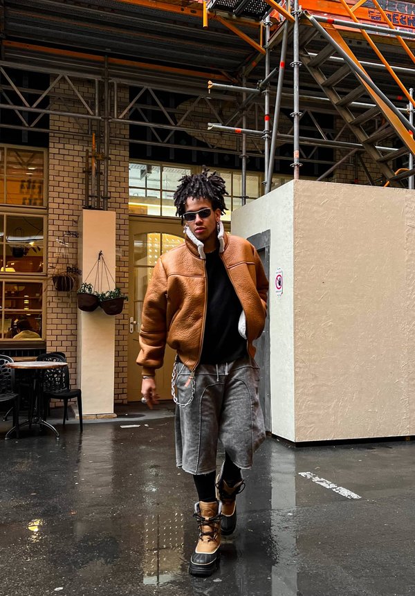 Veste bombardier en cuir marron avec doublure blanche, chemise noire, shorts gris et bottes beiges. Accessoirisée avec des lunettes de soleil et une chaîne. Pavé mouillé.