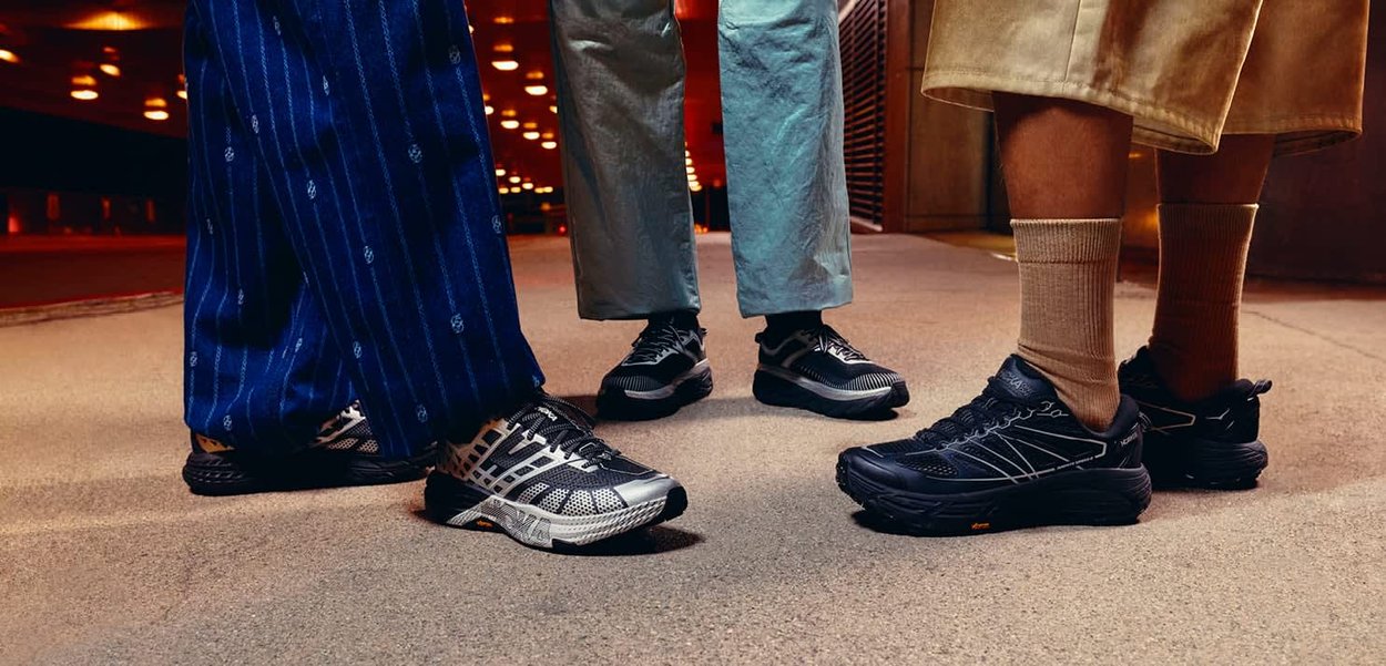 Tre paia di gambe che indossano pantaloni casual e scarpe da ginnastica nere, in piedi su un pavimento tappezzato in un interno poco illuminato.