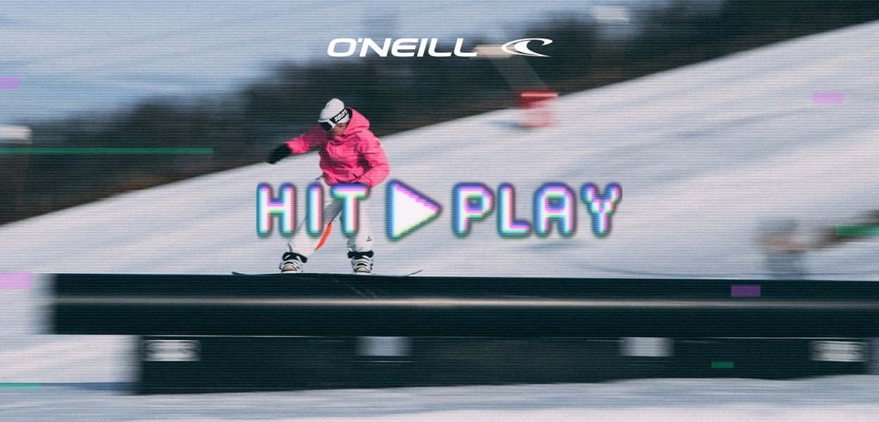 Skifahrer in pinker Jacke und weißer Hose rutscht auf einer Schiene im verschneiten Terrainpark, darüber der Text "HIT PLAY" und das O'Neill-Logo.