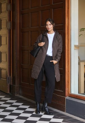 Quiltad brun lång kappa med huva, kombinerad med en vit T-shirt och svarta byxor, ackompanjerad av svarta kängor och en liten handväska.