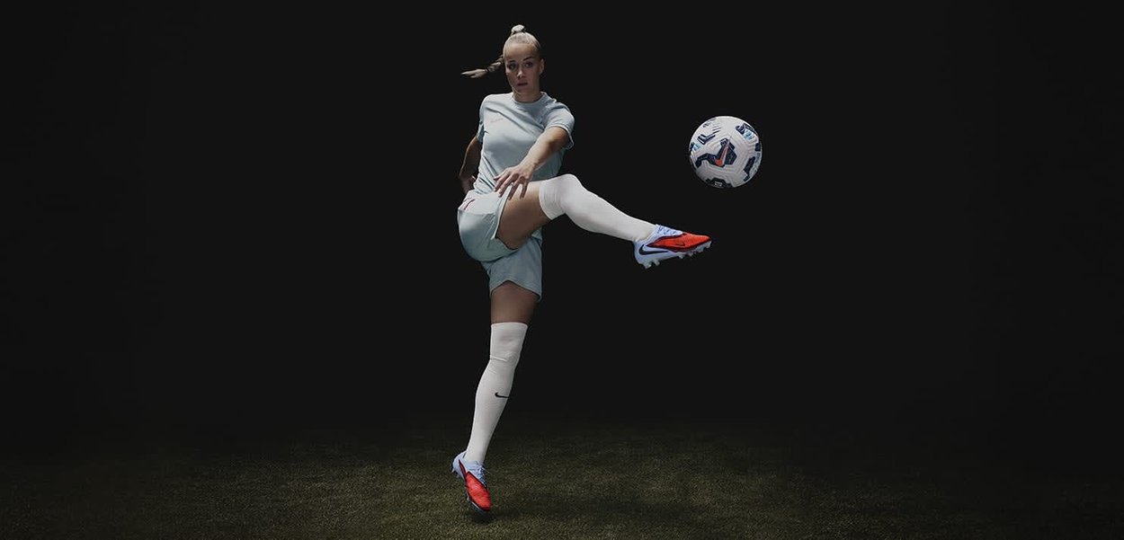 Atleta femenina con camiseta de color azul claro y pantalones cortos blancos, pateando un balón de fútbol blanco, con zapatos de futbol rojos y negros, sobre un fondo oscuro.