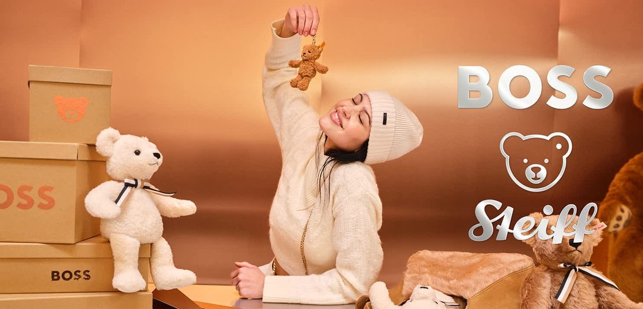 Beige Teddy-Bären, einer mit einem schwarz-weißen Band, platziert neben beigen Boxen mit der Aufschrift "BOSS." Eine Person hält einen kleinen braunen Bären-Schlüsselanhänger.