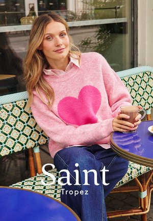 Femme en pull rose avec un grand cœur, jean bleu, assise à une table de café tenant un verre de café, avec le texte Saint-Tropez en dessous.