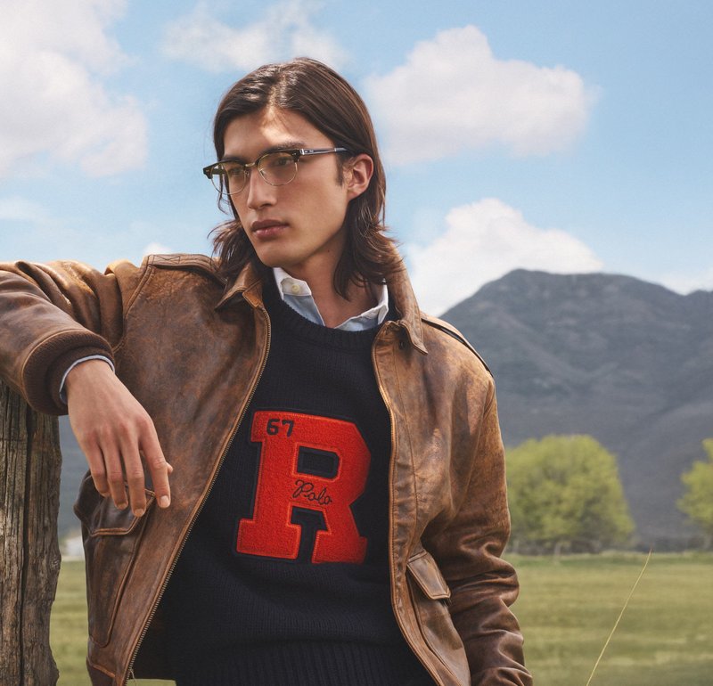 Veste en cuir marron sur un pull marine avec un grand logo rouge "R". Le mannequin porte des lunettes, avec de longs cheveux, devant un paysage naturel.