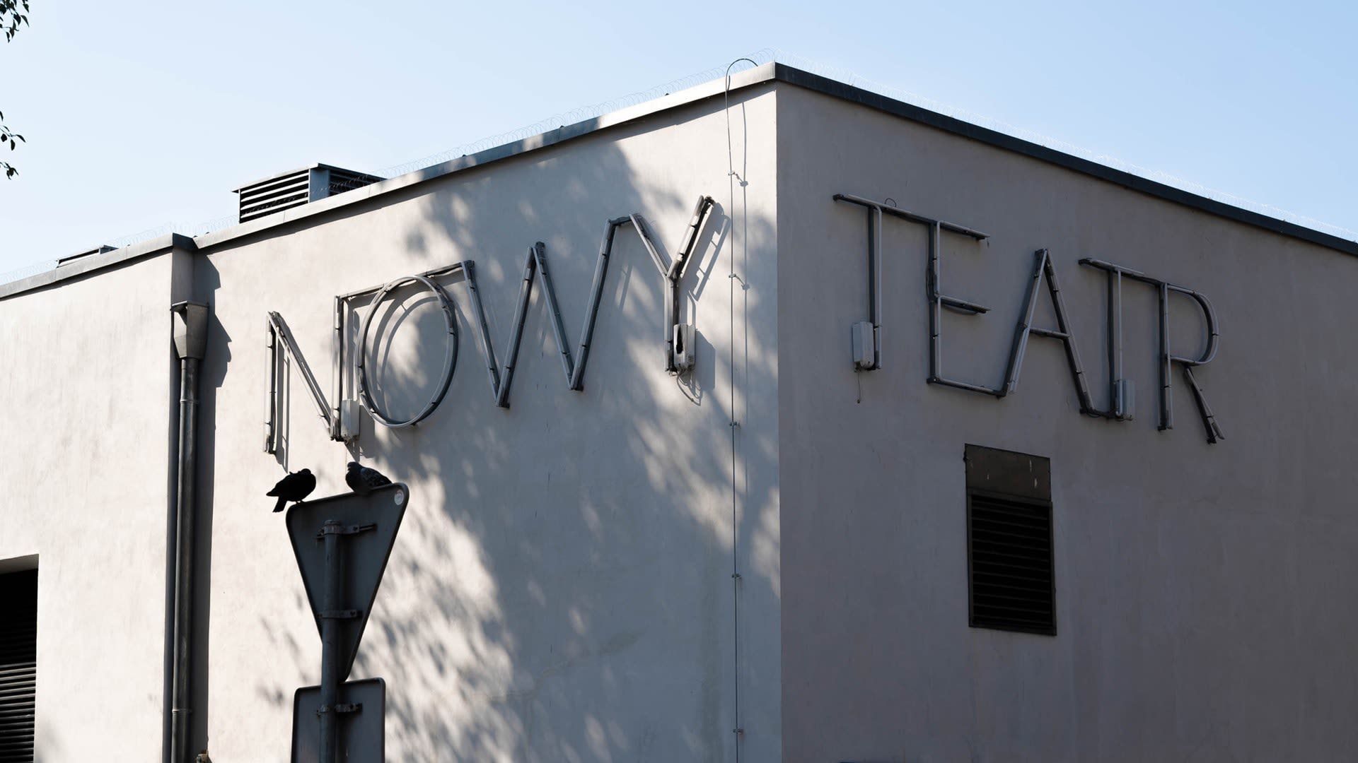 Neon buizenletters die "NOWY TEATR" spellen, gemonteerd op een beige muur, met twee vogels die op een verkeersbord eronder zitten.