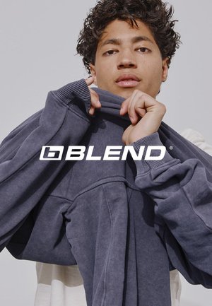 Junger Mann mit lockigem Haar zieht einen dunkelgrauen Pullover über den Kopf vor hellem, einfarbigem Hintergrund, Text "BLEND" in der Mitte.
