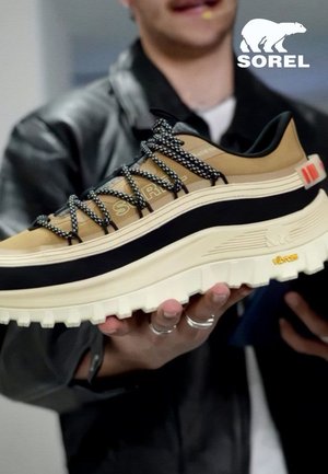 Main tenant une chaussure de sport Sorel marron clair et noire avec une semelle blanche épaisse et des lacets à motif, logo visible en haut à droite.