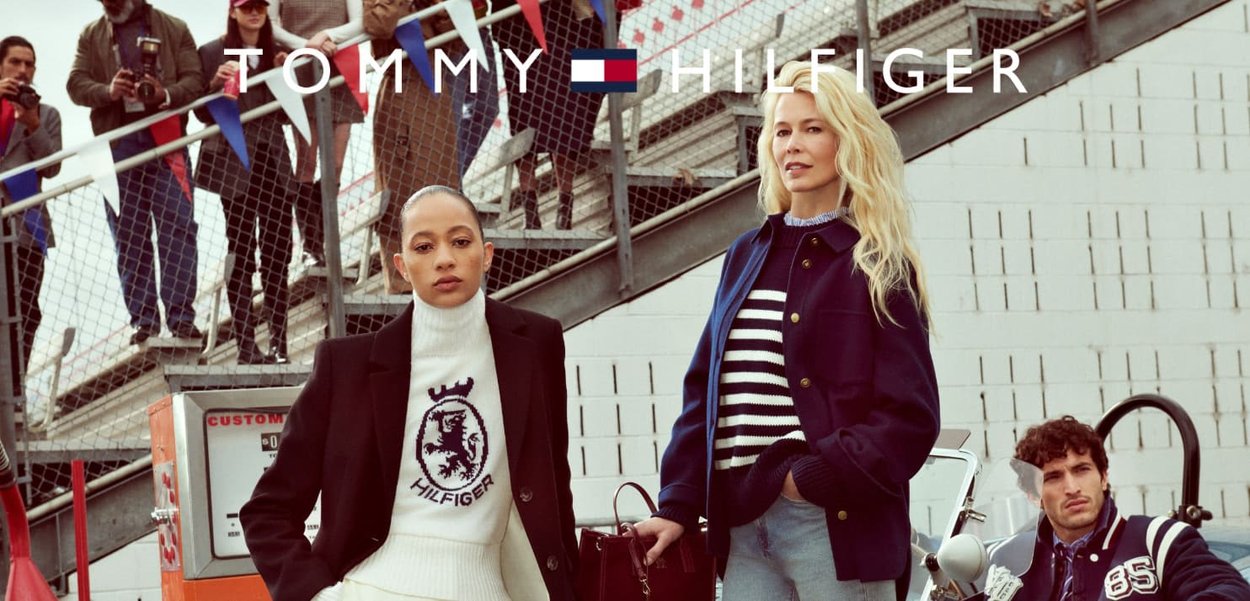 Models wear Tommy Hilfiger apparel: a cream sweater with logo, a navy blazer, and a striped top. Background features spectators and flags.