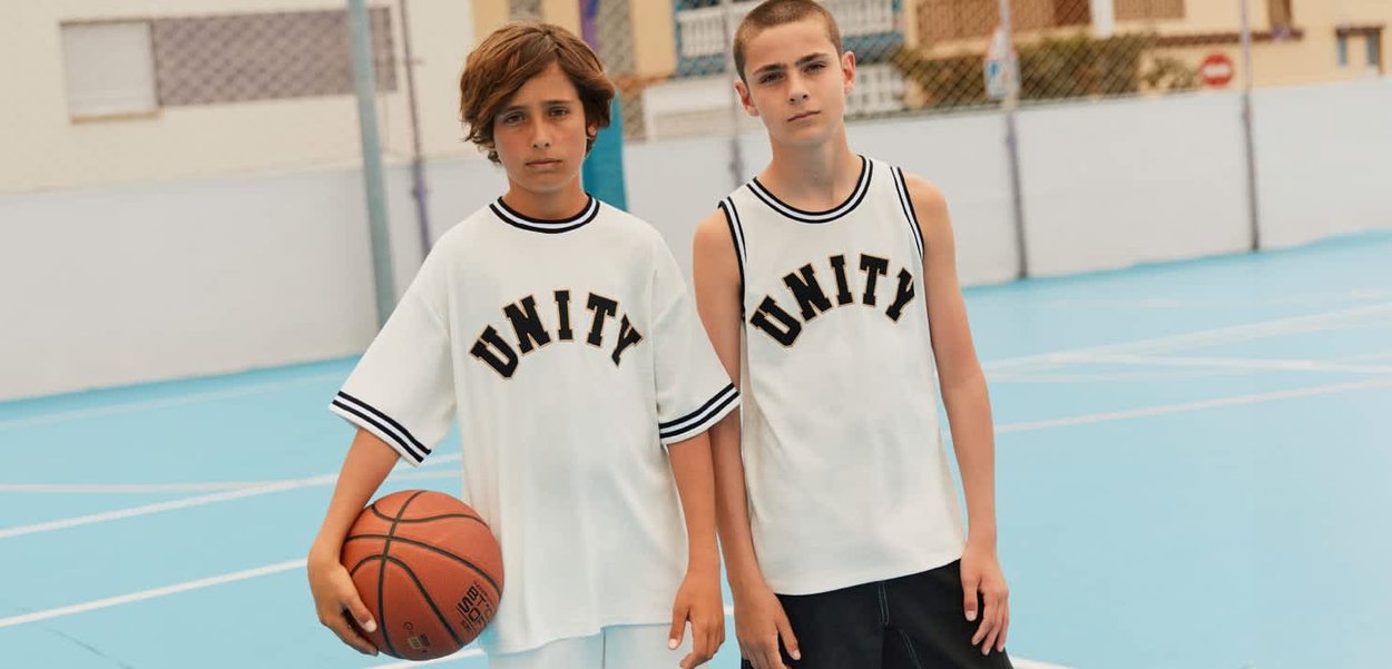 Twee jongens in witte "UNITY" basketbalshirts staan op een buitenveld, waarvan een jongen een basketbal naast zich vasthoudt.