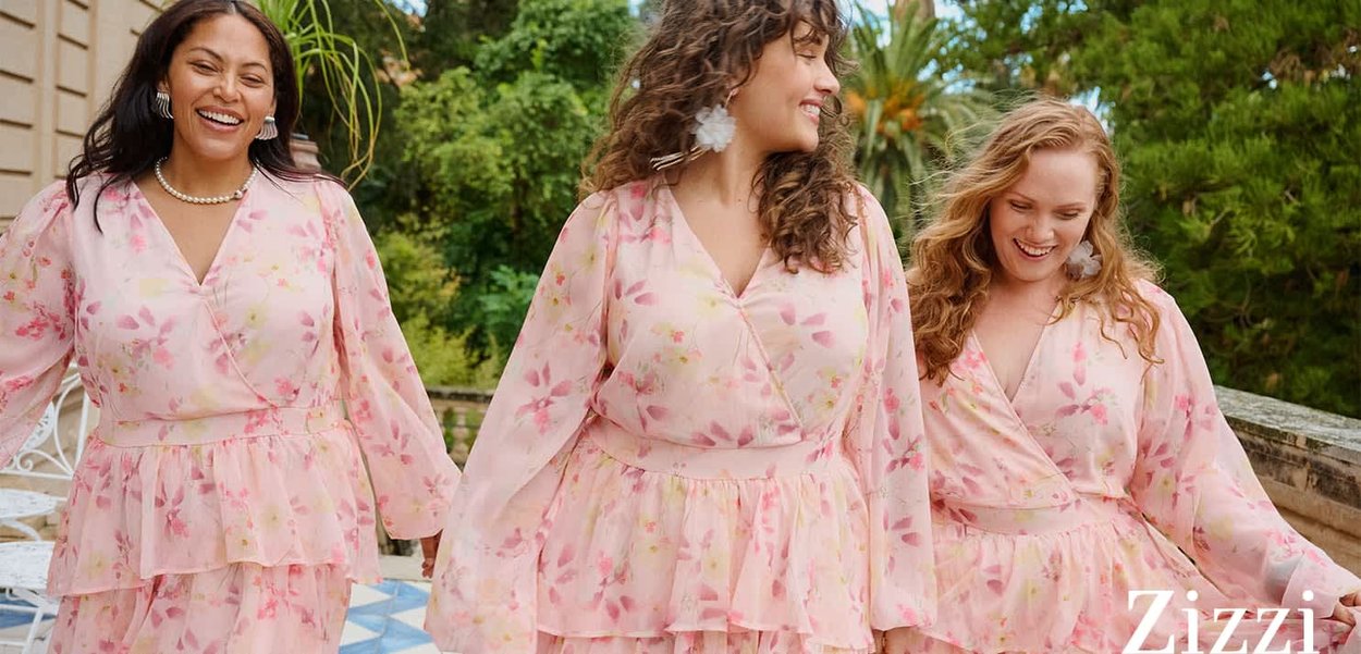 Tre smilende kvinder bærer matchende lyserøde blomstrede kjoler med lagdelte skørter og går udenfor nær grønne planter på en flisebelagt terrasse.