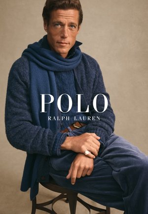 Pull en maille bleu marine avec une écharpe, pantalon en velours côtelé et une bague à la main. Le mannequin est assis sur une chaise devant un fond neutre.