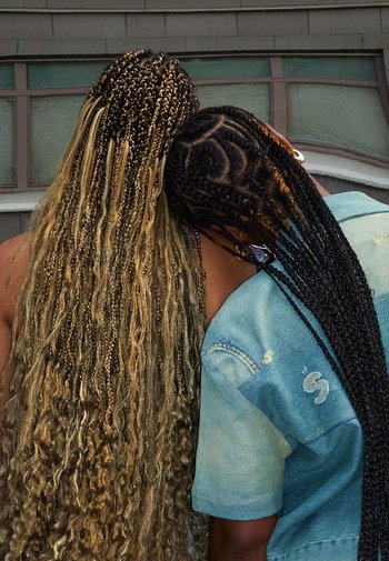 Twee individuen met gevlochten haar, de een lang en golvend in blond en bruin, de ander strak gevlochten in zwart. Beiden dragen spijkerjacks.