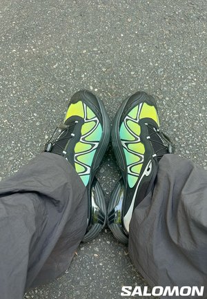 Scarpe da atletica nere e verdi con tomaia in rete, suola curvata e un motivo a onde distintivo. Indossate con pantaloni grigi larghi.