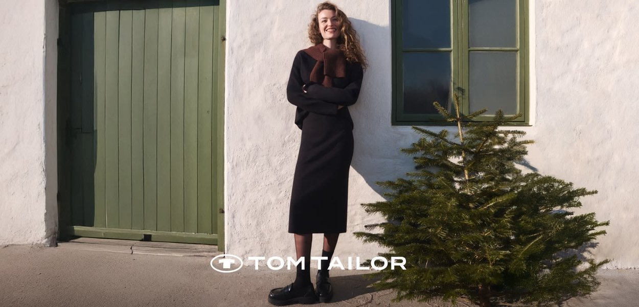 Woman in black dress and brown scarf stands smiling against white wall near green door and window, next to a small pine tree.