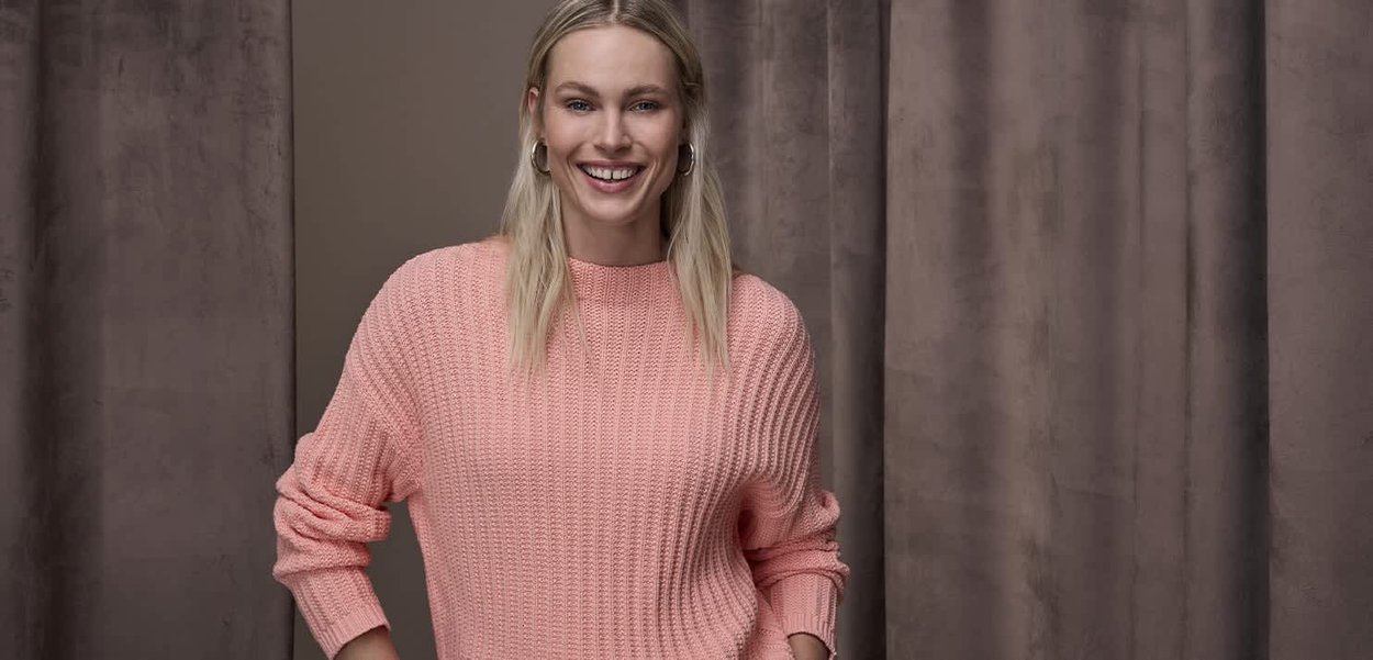 Jasnoróżowy sweter dziany o prążkowanej fakturze i swobodnym kroju, z długimi rękawami i szerokim okrągłym dekoltem na neutralnym tle.