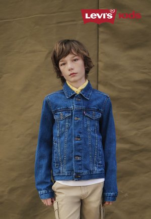 Spijkerjack met een klassieke snit in blauw, voorzien van een knoopsluiting, twee borstzakken en een zachte textuur. Draagt over een geel shirt.