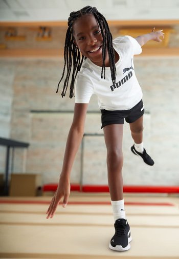 Niño con una camiseta blanca de Puma, pantalones cortos negros y zapatillas negras, equilibrándose en un pie en un gimnasio con suelo de madera.