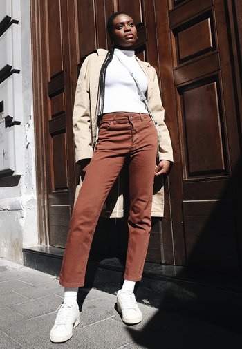 Pantalons marron taille haute, col roulé blanc, manteau beige, avec des baskets blanches. Debout contre une porte en bois au soleil.