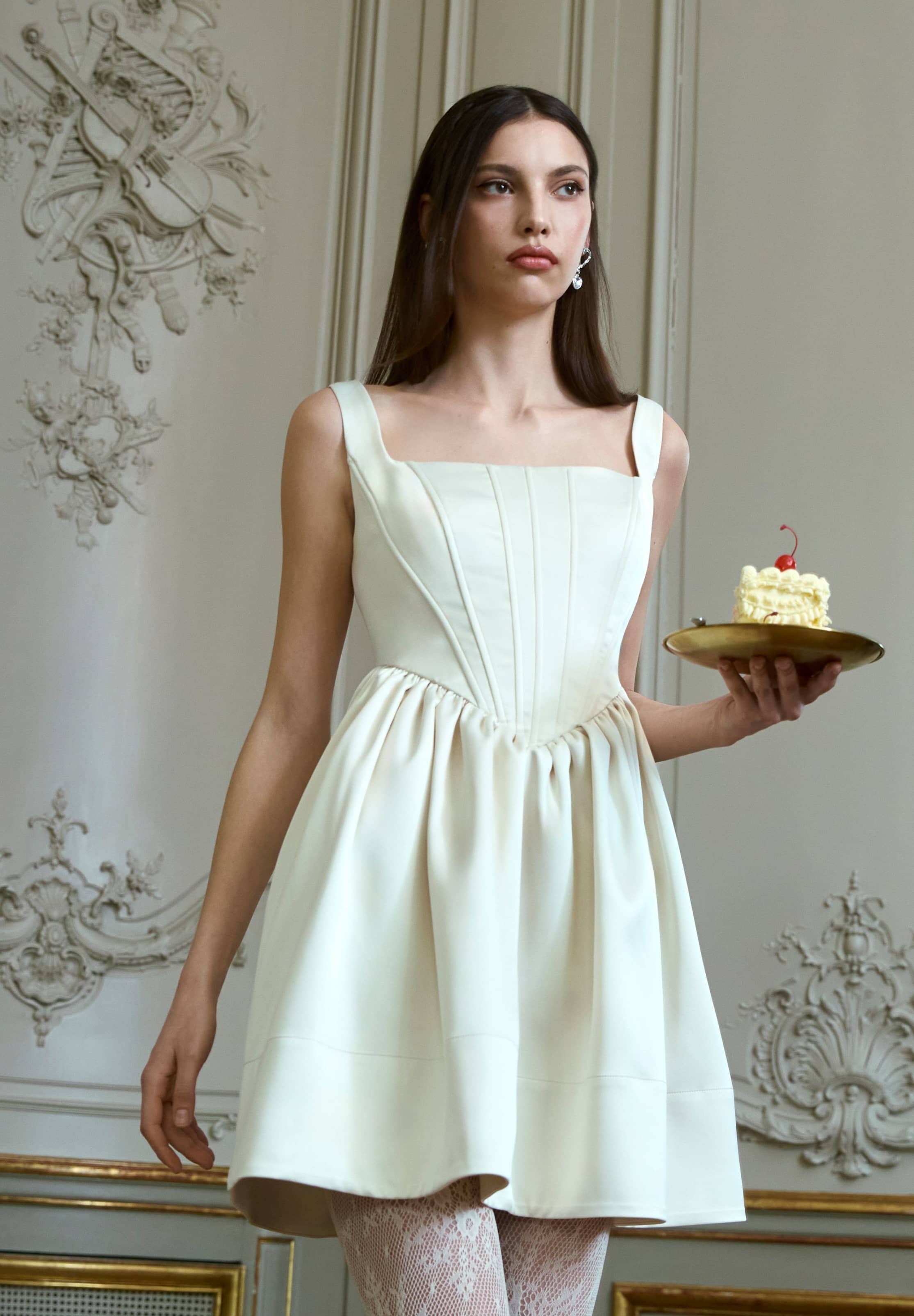 Cremfarbendes ärmelloses Kleid mit quadratischem Ausschnitt, plissiertem Oberteil, ausgestelltem Rock und filigranen Spitzentights; Modell hält einen Kuchen auf einem Teller.