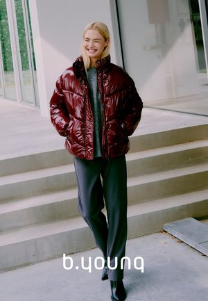 Giacca puffer bordeaux con una finitura lucida, colletto alto, indossata sopra un maglione grigio, abbinata a pantaloni neri e stivaletti.