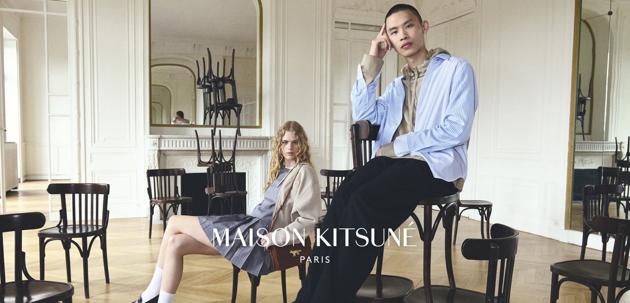 Zwei Modelle sitzen auf dunklen Holzstühlen in einem hellen, eleganten Raum, in dem gestapelte Stühle in einem großen Wandspiegel reflektiert werden.