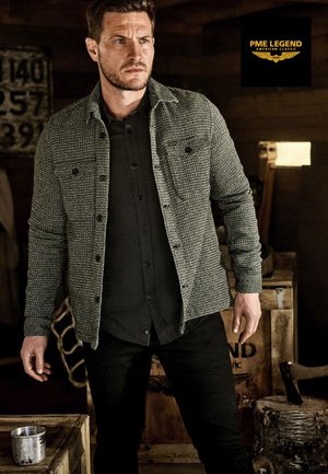 Grau gewebte Hemdjacke mit einem strukturierten Muster, schwarzen Knöpfen und zwei Brusttaschen. Kombiniert mit einem schwarzen Shirt und dunklen Jeans.