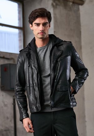 Giacca da motociclista in pelle nera con cerniere, felpa con cappuccio sotto, maglia grigia lavorata a trama e dettagli discreti sulle tasche.