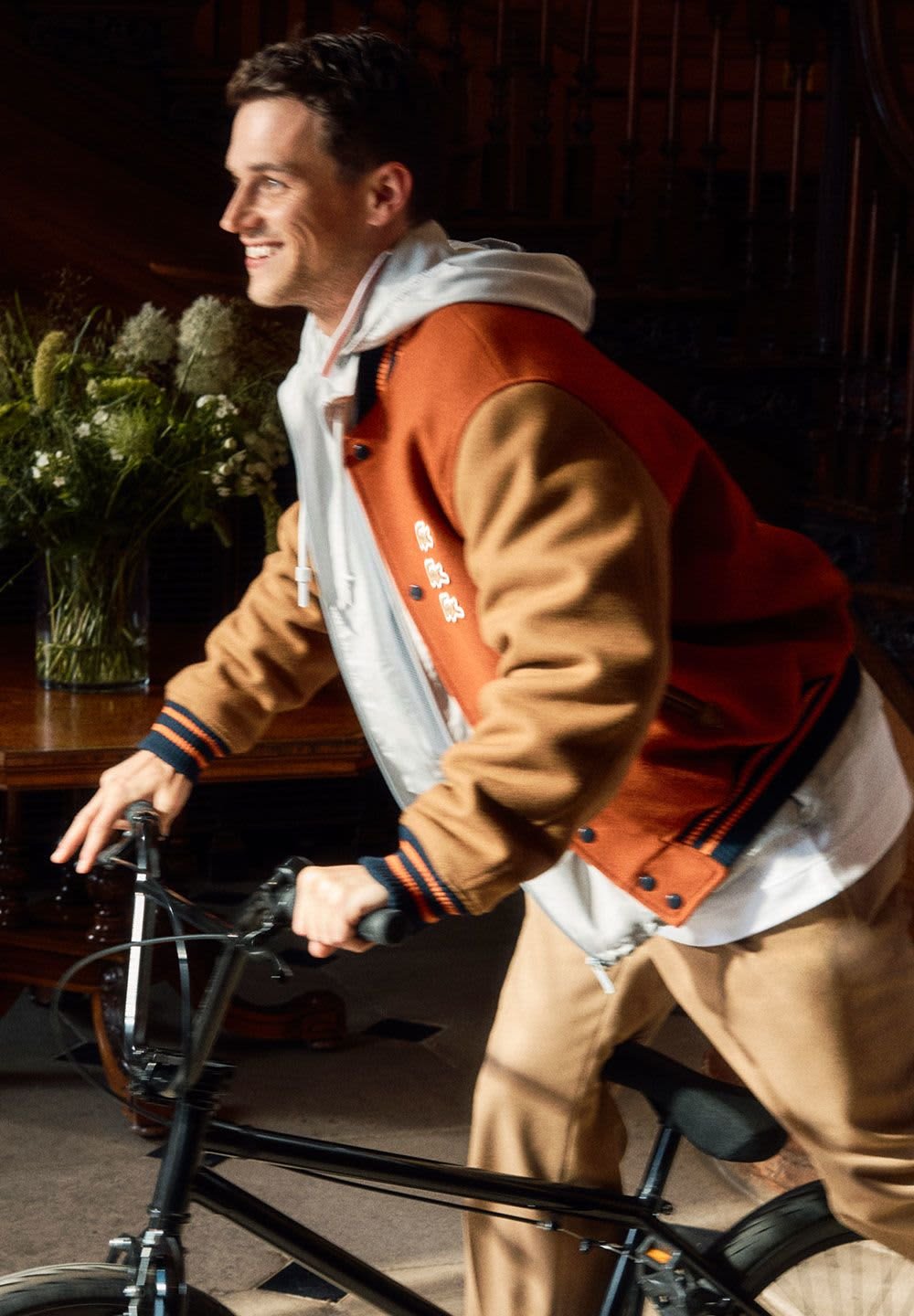 Veste de varsity orange et marron avec des accents noirs, portée sur un sweat à capuche clair. Un cavalier tenant un vélo noir, entouré de verdure.