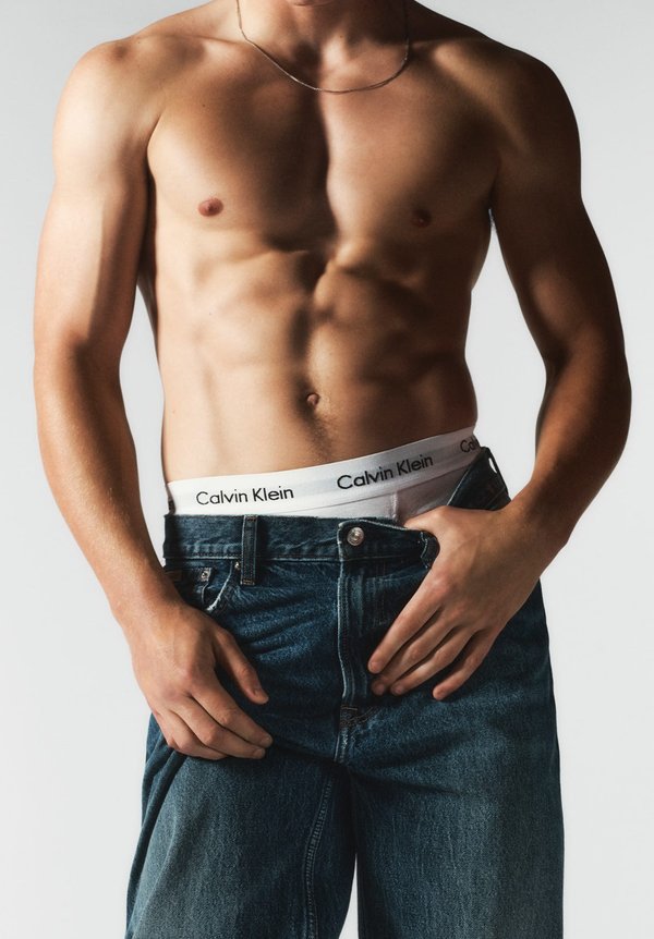 Męski tors noszący srebrny łańcuszek, z ciemnoniebieskimi dżinsami i białym paskiem Calvin Klein z czarnym logo. Gładka faktura skóry.