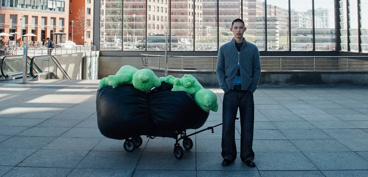 Black fabric cart with green plush figures inside, standing on tiled pavement. Person in gray cardigan and dark trousers next to it.