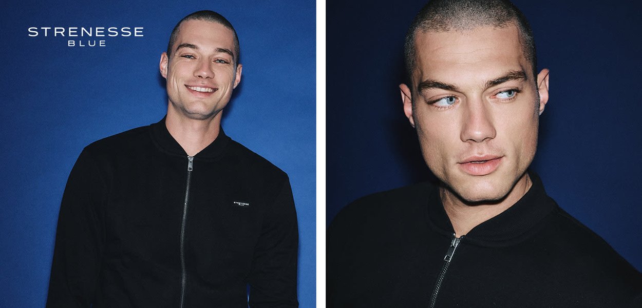 Junger Mann mit kurzem Haarschnitt, der eine schwarze Jacke mit Reißverschluss trägt, posiert lächelnd und seitlich blickend vor einem dunkelblauen Hintergrund.