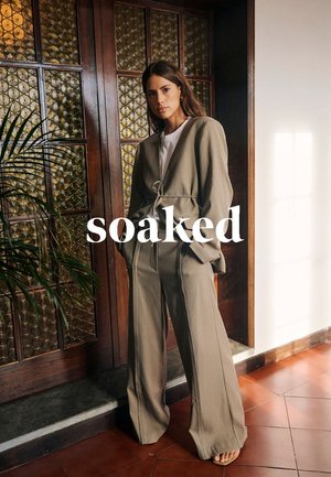 Khakifarbenes zweiteiliges Outfit mit weit geschnittenen Hosen und einem taillierten, mit Gürtel versehenen Sakko. Der Stoff wirkt glatt und hat eine maßgeschneiderte Passform. Natürlicher Hintergrund mit Holzakzenten.