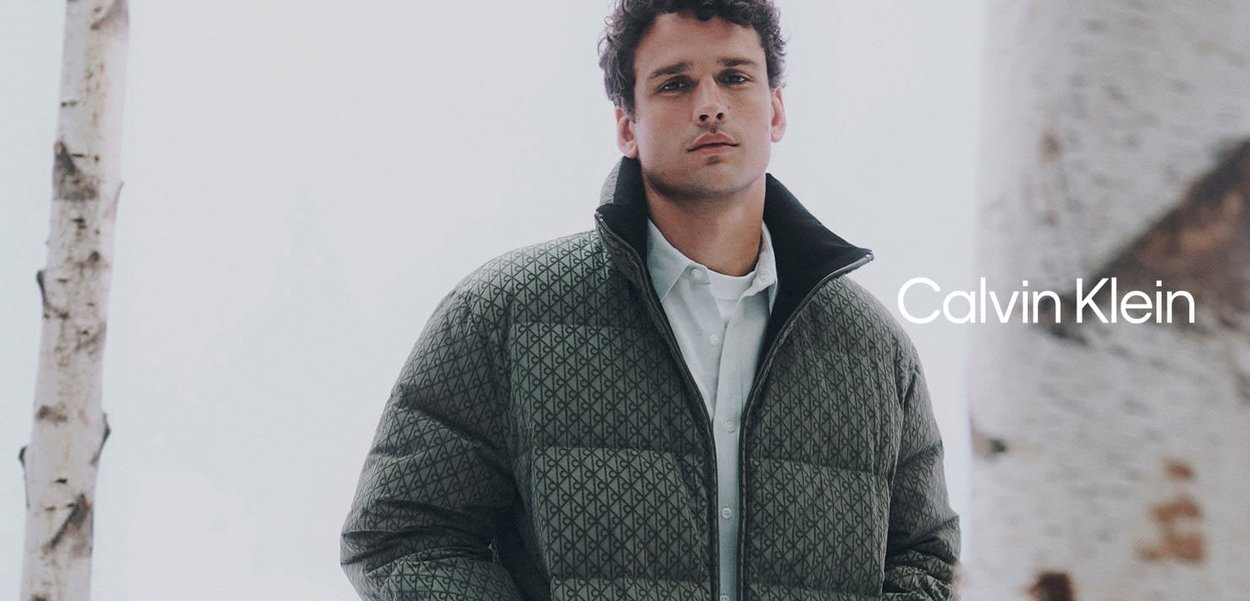 Chaqueta acolchada verde con un patrón de diamantes, cuello alto y cierre de cremallera. Llevada sobre una camisa blanca. Fondo con abedules desenfocados.