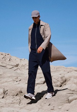 Man loopt op rotsachtig terrein, gekleed in een beige trenchcoat, marineblauw rits-outfit, grijze pet en witte sneakers onder een heldere blauwe lucht.