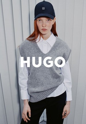 Pull sans manches gris tricoté porté sur une chemise blanche à col, accompagné d'un pantalon noir. Une casquette foncée arbore un logo. Le fond présente des rayures verticales.