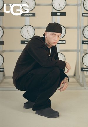 Homme en tenue noire et bonnet accroupi portant des bottes grises UGG, avec plusieurs horloges affichant des villes du monde sur le mur derrière lui.