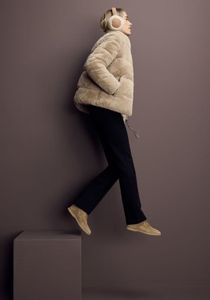 Beżowa kurtka z sztucznego futra w poziome paski, czarne spodnie i beżowe zamszowe buty. Model nosi jasnobeżowe słuchawki, na tle ciemnej ściany.