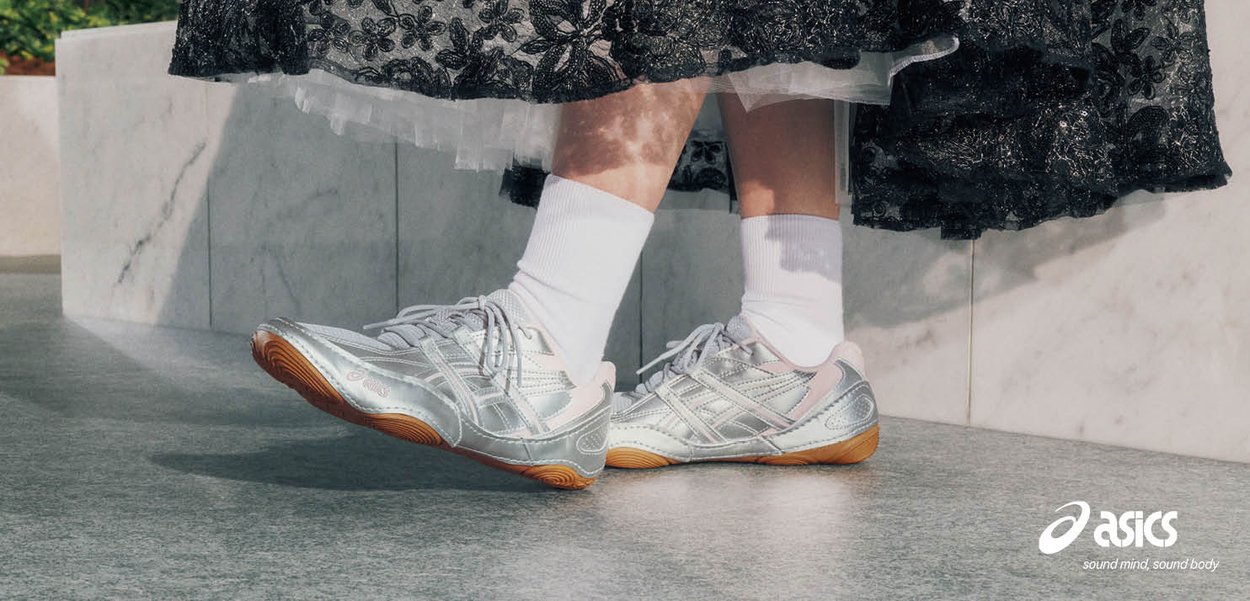 Beine mit weißen Socken und silbernen Asics-Sneakern kombiniert mit einem schwarzen Spitzenrock, stehend auf einem grauen Boden nahe einer Marmorwand.