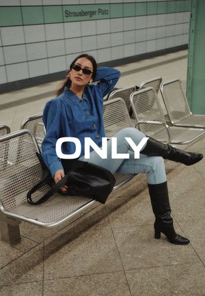 Donna con camicia di jeans blu, jeans chiari e stivali neri seduta su panchine metalliche nella stazione della metropolitana di Strausberger Platz con una borsa nera in mano.