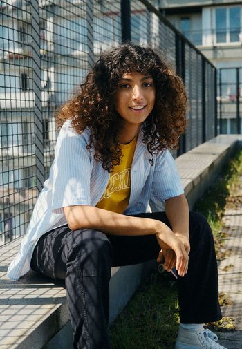Chemise à rayures à manches courtes, t-shirt jaune avec texte, pantalons sombres et baskets blanches. Cheveux bouclés, assis sur un chemin près d'une clôture.