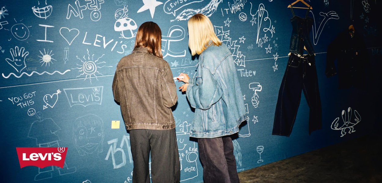 Denimjacken in dunkelblau und hellblau, getragen von zwei Personen, vor einer blauen Wand, die mit Kritzeleien und dem prominent platzierten Levi's-Logo bedeckt ist.